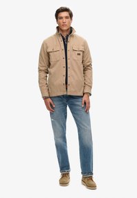 Beige canvas jacket met drukknoopjes en twee borstzakken, gedragen over een marineblauw shirt. Lichtblauwe distressed jeans en beige schoenen maken de look compleet.