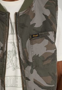 Chaleco de camuflaje con cremallera frontal, cuello acanalado verde oliva y un bolsillo en el pecho con el logo de Wrangler. Tejido texturizado y diseño ajustado.