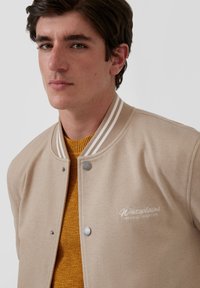 Beżowa kurtka typu varsity z szarym prążkowanym kołnierzem, srebrnymi napami i wyszywanym logo "Whiteplains"; noszona na musztardowym swetrze.