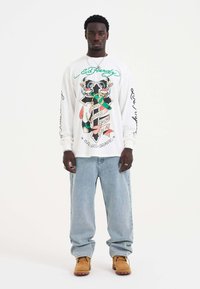Witte lange mouwen shirt met kleurrijk grafisch ontwerp, vergezeld van lichtblauwe jeans en tan kleurige laarzen. Ontspannen pasvorm, met een opvallende print aan de voorkant.