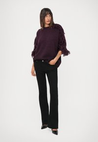Žena oblečená v purpurovom svetríku s franjami, s čiernymi priliehavými bootcut nohavicami a čiernymi špicatými lodičkami, stojí s jednou rukou vo vrecku na bielom pozadí.