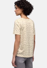 Nainen, jolla on kiharat hiukset ja joka pukeutunut beigeen, lyhythihaiseen paitaan, jossa on vaaleahiirileopardi-kuosi, sekä harmaisiin farkkuihin, katsoo vasemmalle valkoista taustaa vasten.