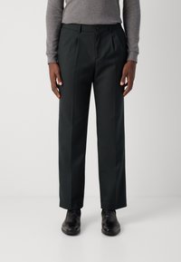 Pantalon noir avec une coupe droite, texture lisse, plis à l'avant et fermeture à bouton. Associé à des chaussures noires et un haut gris.