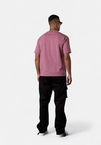 Mann mit Nackentattoo und Sonnenbrille, der ein staubiges rosa T-Shirt, schwarze Hosen und weiße Sneaker trägt und sich mit dem Rücken zu einer einfarbigen Hintergrund steht.