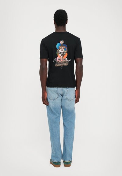 Man wearing black T-shirt with colorful snake and skull graphic and light blue jeans, standing facing away against plain white background.