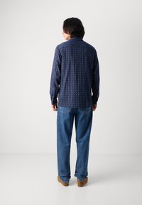 Camisa de rayas navy con rayas verticales, de manga larga y con puños abotonados, combinada con jeans azules holgados y zapatos marrones.