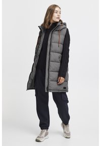 Ärmellose, graue Daunenweste mit Kapuze, ausgestattet mit schwarzem Rand und orangefarbenen Kordeln. Getragen über einem dunklen Pullover und kombiniert mit marineblauen Hosen.