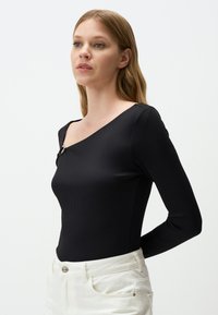 Zwart geribbeld langetermijn top met een asymmetrische halslijn en een decoratieve gouden knoopaccent aan de linkerschouder. Gepaard met witte broeken.