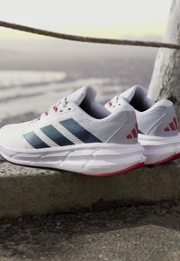 Baskets de course blanches en mesh et matériaux synthétiques. Présentent des accents bleus et rouges, avec un logo à trois rayures parallèles sur le côté.