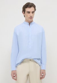 Giovane uomo che indossa una camicia a collo alla mandarina blu chiaro con bottoni bianchi e pantaloni beige, su uno sfondo chiaro e uniforme.