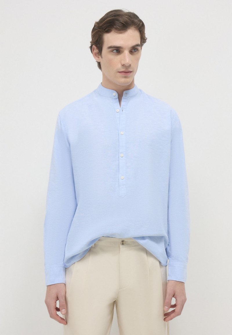 Giovane uomo che indossa una camicia a collo alla mandarina blu chiaro con bottoni bianchi e pantaloni beige, su uno sfondo chiaro e uniforme.