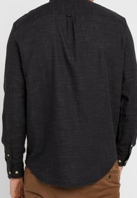 Tumman sävyinen pitkähihainen paita, jossa on teksturoidusta kankaasta valmistettu selän laskos, napilliset kalvosimet ja kaartuva helma.