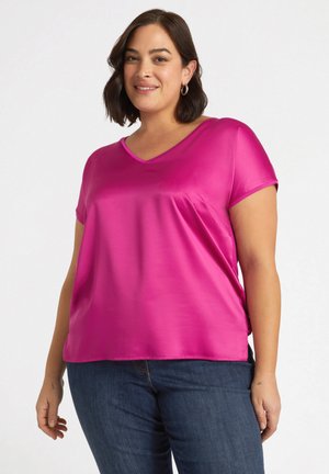 Haut à manches courtes en satin rose vif avec un décolleté en V. Silhouette fluide avec une texture lisse et des fentes sur les côtés. Porté avec un jean bleu.