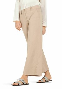 Beige wide-leg trousers made from smooth fabric, featuring a high waist and front pockets. Paired with flat, metallic slide sandals.