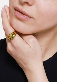 Gouden ring met een gladde, glanzende oppervlakte, gedragen aan de rechterhand en leunend tegen een kin. De huid lijkt natuurlijk en lichtelijk textuur.