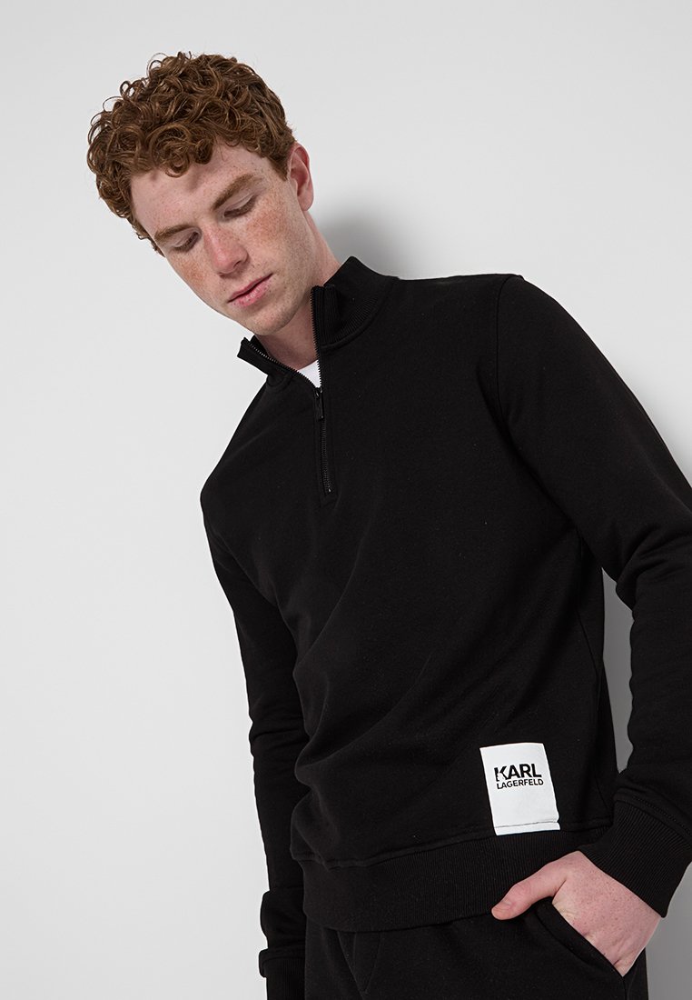 Junger Mann mit lockigem roten Haar, der einen schwarzen Sweatshirt mit Viertelreißverschluss trägt, das am Saum ein weißes Karl Lagerfeld-Logo-Patch aufweist.