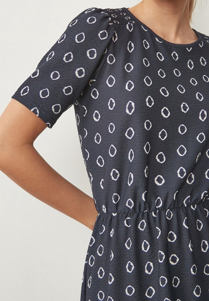 Robe bleu marine avec motif de cercles blancs irréguliers, manches bouffantes courtes et taille élastique froncée portée par un mannequin.