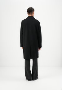 Abrigo de lana negro con cuello de muesca, corte recto y mangas largas, combinado con pantalones de campana gris y zapatos negros.
