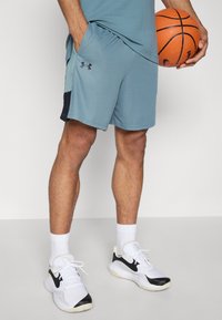 Mann in blauen Sportshorts und weißen Turnschuhen, der einen orangefarbenen Basketball neben sich hält, vor einfarbigem Hintergrund.