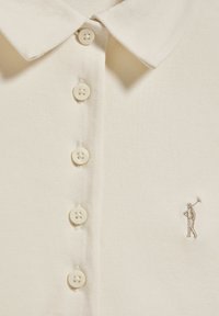 Krämfärgad polo med texturerad tyg, med klassisk krage, fem knappar och en liten broderad golfare-logotyp på vänster sida.