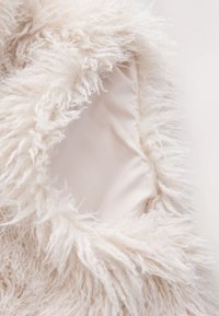 Flauschiger weißer Stoff mit langen, dicken Fasern am Rand, der eine glatte, hellbeige Innenseite zeigt. Weiches und strukturiertes Erscheinungsbild.