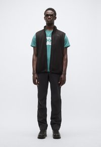 Μαύρο αμάνικο γιλέκο πάνω από τουρκουάζ μπλουζάκι με λευκό λογότυπο, σε συνδυασμό με μαύρο παντελόνι και μαύρα παπούτσια· το μοντέλο φοράει γυαλιά ηλίου.