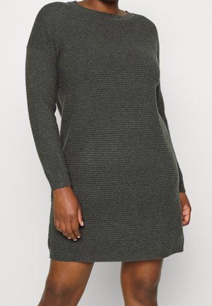 Person iført en ribstrikket mørkegrå langærmet kjole med rund hals, stående mod en ensartet lys baggrund.