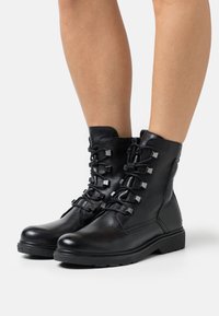 Botas de tobillo de cuero negro con punta redonda, cierre de cremallera y acentos decorativos de hardware cuadrado en los cordones. Suela de goma para tracción.