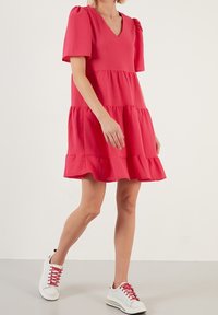 Růžové vrstvené šaty s výstřihem do V a krátkými rozevlátými rukávy. Spárováno s bílými teniskami s červenými tkaničkami. Hladká látka, ležérní střih.