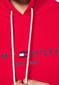 Felpa rossa con cappuccio, cordini bianchi e ricamo "TOMMY HILFIGER EST. 1985" con logo sul petto, mano che regge un cordino.