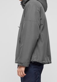 Grå huvtröja med en slät, matt tyg; har justerbara snörningar, sidofickor och kardborremuddar. Enkelt, funktionellt design.