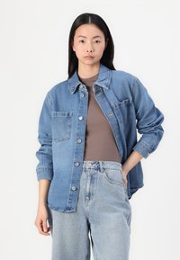 Jaqueta de denim azul claro com botões de pressão, dois bolsos no peito e um corte relaxado, usada sobre uma blusa castanha e combinada com jeans azul claro.