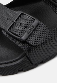 Melnas sandaļi ar tekstūru gumijas virsmu, aprīkoti ar divām regulējāmām siksnām, kas fiksētas ar melniem plastmasas sprādzēm. Vieglas konstrukcijas.