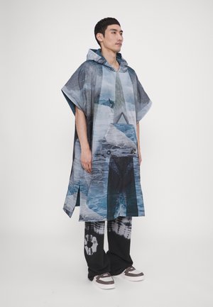 Hooded poncho with a photographic print of shades of blue and gray, featuring a front pocket. Worn with black tie-dye pants and sneakers.