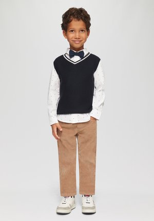 Navy gebreide vest over een wit overhemd met stippen, bruine corduroy broek en witte sneakers. De outfit heeft een vlinderdas en een casual pasvorm.