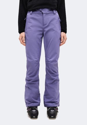 Un mannequin portant un pantalon de ski violet, un haut noir et des chaussures de ski noires se tient devant un fond uni.