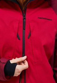 Rode waterdichte jas met zwarte ritsen en verstelbare kenmerken. De stof is glad met een matte afwerking. Het merklogo is zichtbaar op de borst.