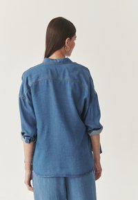 Jeans košulja u srednje plavoj boji, opušteni kroj, dugi rukavi s roliranim manžetama, detalj na leđima, kontrastni šavovi, glatka tekstura.