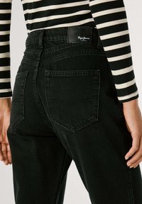 Melna džinsu auduma bikses ar augstu jostasvietu, kurām ir divas aizmugurējās kabatas un logotipa uzlīme jostasvietā. Audumam ir smalka tekstūra.