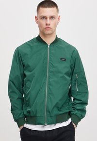 Grønn bomberjakke laget av glatt nylon. Har en front glidelås, ribbede mansjetter og en sidelomme med glidelås. Enkel design uten mønstre.