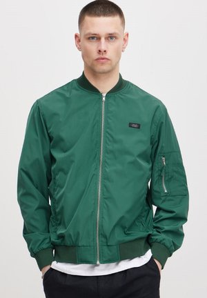 Grønn bomberjakke laget av glatt nylon. Har en front glidelås, ribbede mansjetter og en sidelomme med glidelås. Enkel design uten mønstre.