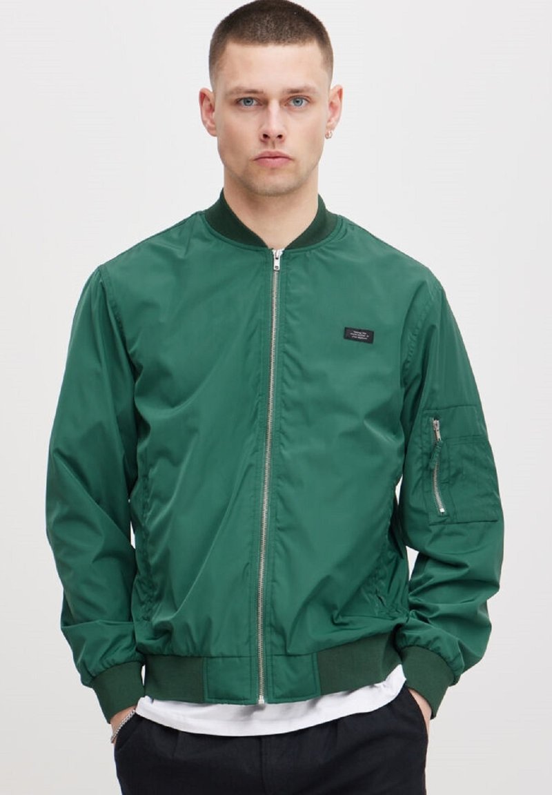 Grønn bomberjakke laget av glatt nylon. Har en front glidelås, ribbede mansjetter og en sidelomme med glidelås. Enkel design uten mønstre.