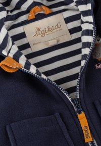 Marineblaue Fleecejacke mit gestreiftem Innenfutter, orangefarbener Reißverschlussöse und einem kleinen gestickten Detail auf der Brusttasche.