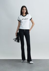 Jeune femme debout portant un t-shirt blanc avec le texte "OFF-white", un pantalon noir, des chaussures argentées à bout pointu, tenant une pochette noire à franges sur un fond uni.