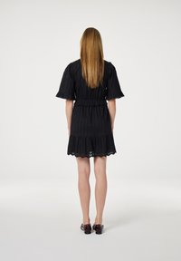 Zwarte jurk met korte, opgeblazen mouwen, gestructureerde stof, elastische taille en golvende zoom. G gedragen met donkere schoenen, gezien van achteren.