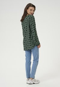 Chemise à manches longues en motif de losanges noir et vert, associée à un jean bleu clair et des baskets grises, présentant une coupe décontractée et une texture douce.