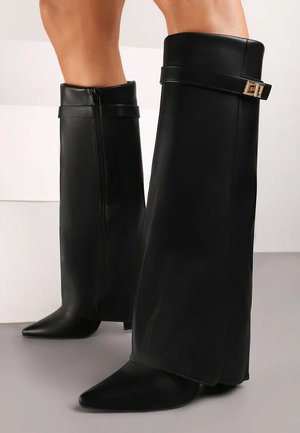 Jambes vêtues de bottes hautes en cuir noir avec fermeture éclair latérale et sangle décorative avec boucle dorée près du haut.