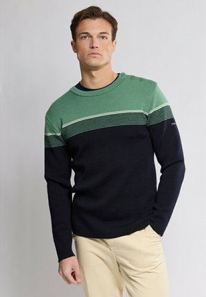 Homme portant un pull rayé vert et noir avec des boutons sur l'épaule et un pantalon beige, debout devant un fond uni.
