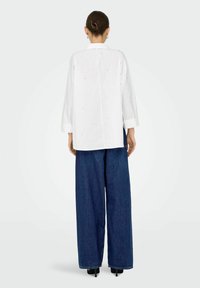 Camicia bianca, oversize, con maniche lunghe, caratterizzata da sottili decorazioni, abbinata a pantaloni blu in denim a gamba larga e tacchi neri.