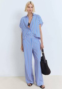 Camicia oversize a righe blu e bianche con maniche corte, abbinata a pantaloni larghi coordinati. Una borsa intrecciata nera completa l'outfit.